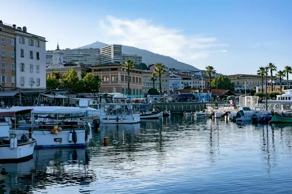 Peaceful harbor in Ajaccio, Corsica with moored boats reflecting cityscape and mountain backdrop.
