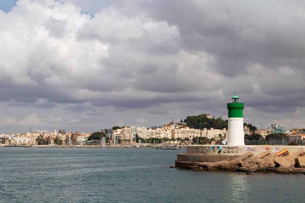 Scenic view of Cartagena's coastline with a green lighthouse and city backdrop under cloudy skies.