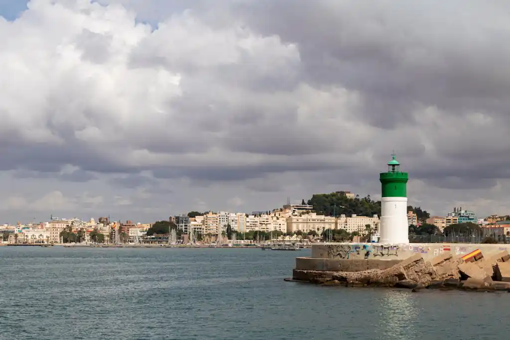 Scenic view of Cartagena's coastline with a green lighthouse and city backdrop under cloudy skies.